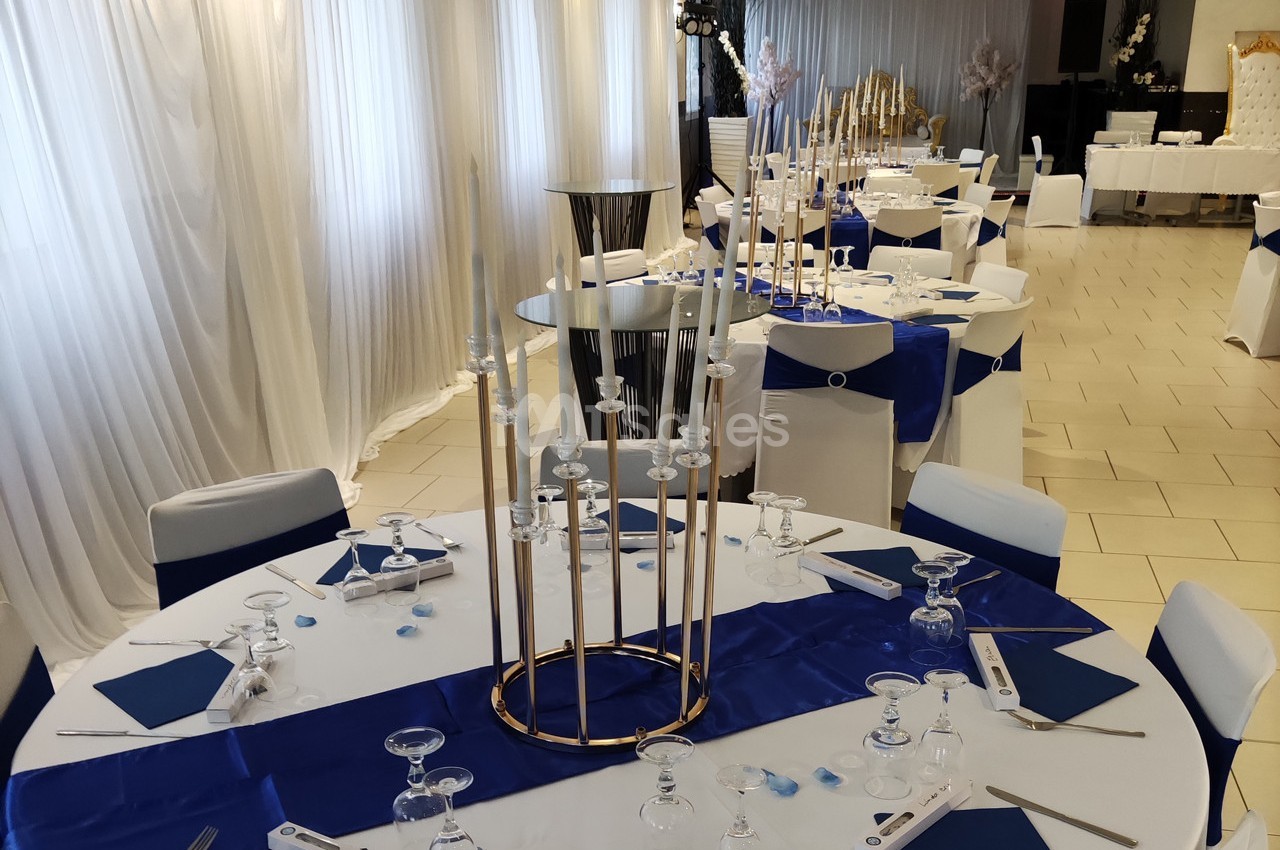 Salle de réception décorée avec des tables rondes, nappes blanches et bleues, et chandeliers dorés au centre.