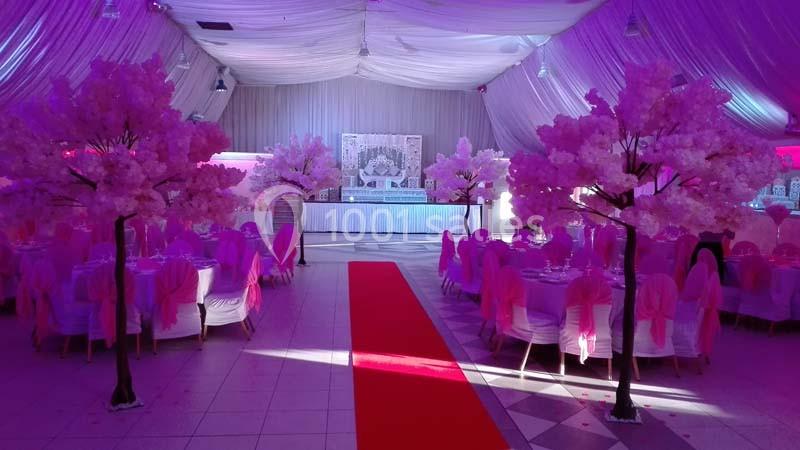 Salle de réception décorée de rose avec tables dressées, arbres artificiels fleuris et tapis rouge central.