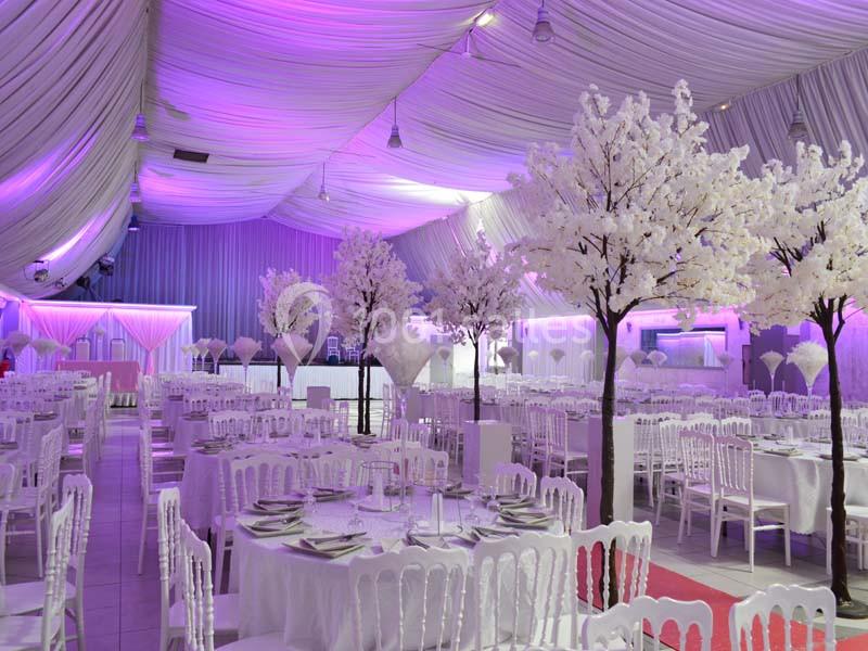 Salle de réception décorée avec des tables rondes, des chaises blanches et des arbres artificiels fleuris sous un éclairage…