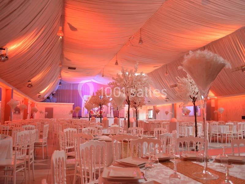 Salle de réception décorée avec des tables dressées, des chaises blanches et des arbres artificiels sous un plafond drapé.
