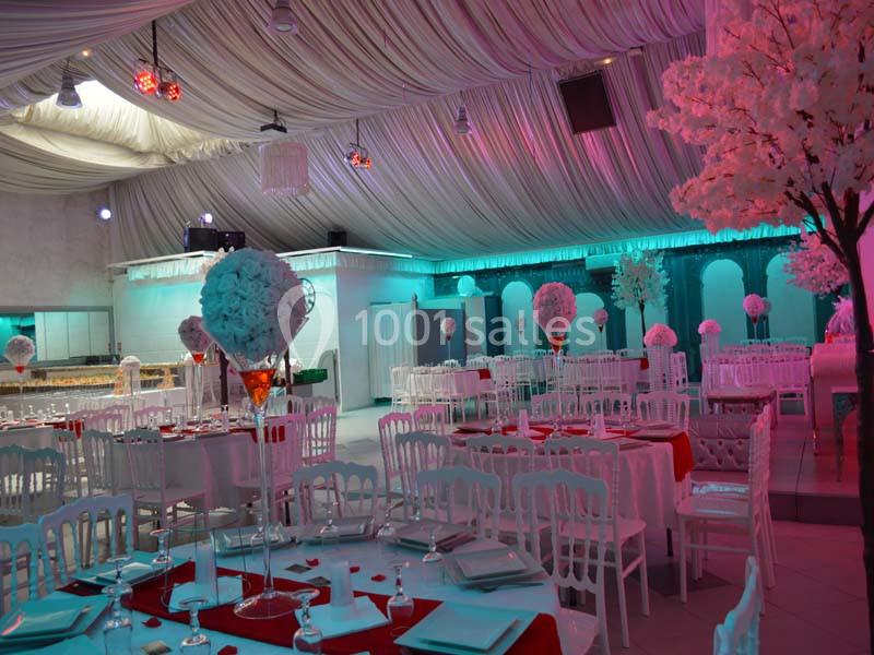 Salle de réception décorée avec des tables dressées, des chaises blanches et des centres de table floraux sous un éclairage…