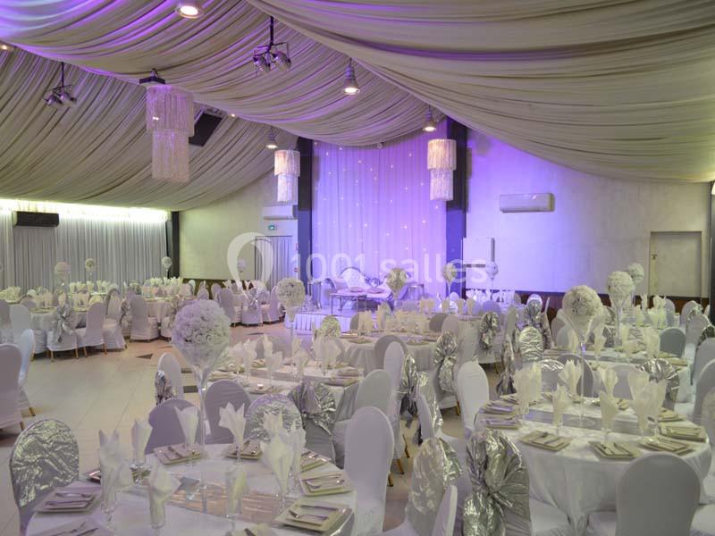Salle de réception décorée avec des tables rondes, nappes blanches, chaises couvertes et éclairage violet tamisé.