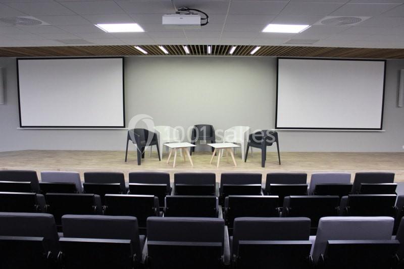 Salle de conférence vide avec des rangées de sièges, deux écrans de projection et trois chaises autour de petites tables.