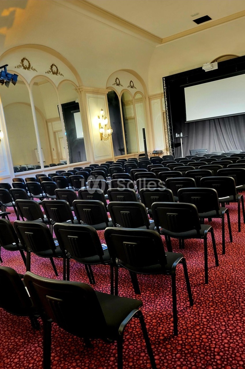 Salle de conférence avec rangées de chaises noires alignées, écran de projection et miroirs sur les murs.