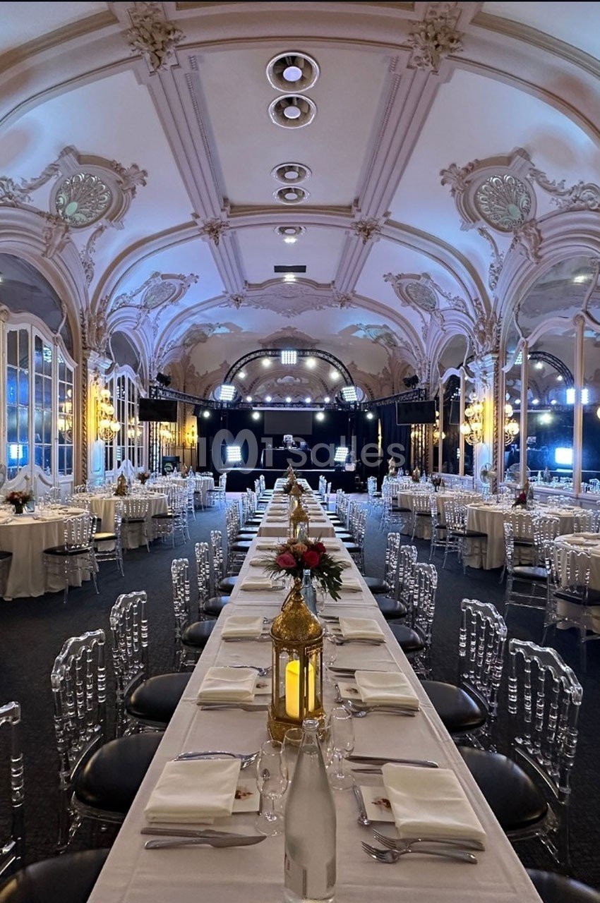 Salle de réception élégante avec tables dressées, chaises transparentes et décoration florale sous un plafond orné.