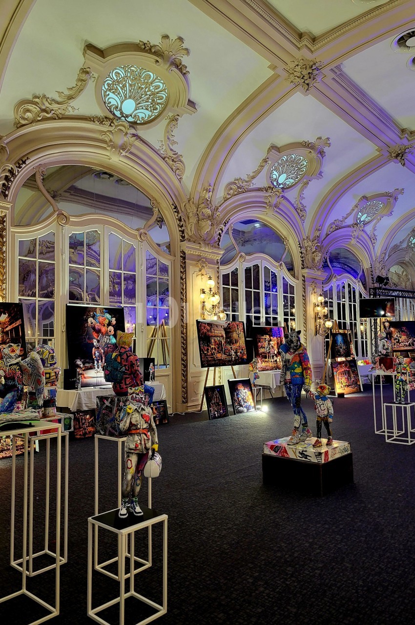 Salle ornée de moulures et vitraux, exposant des sculptures colorées sur des socles et des tableaux lumineux.