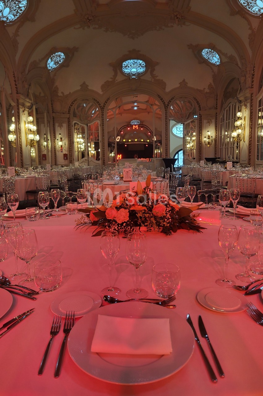 Salle de réception élégante avec tables dressées, nappes blanches, vaisselle raffinée et éclairage tamisé rouge.