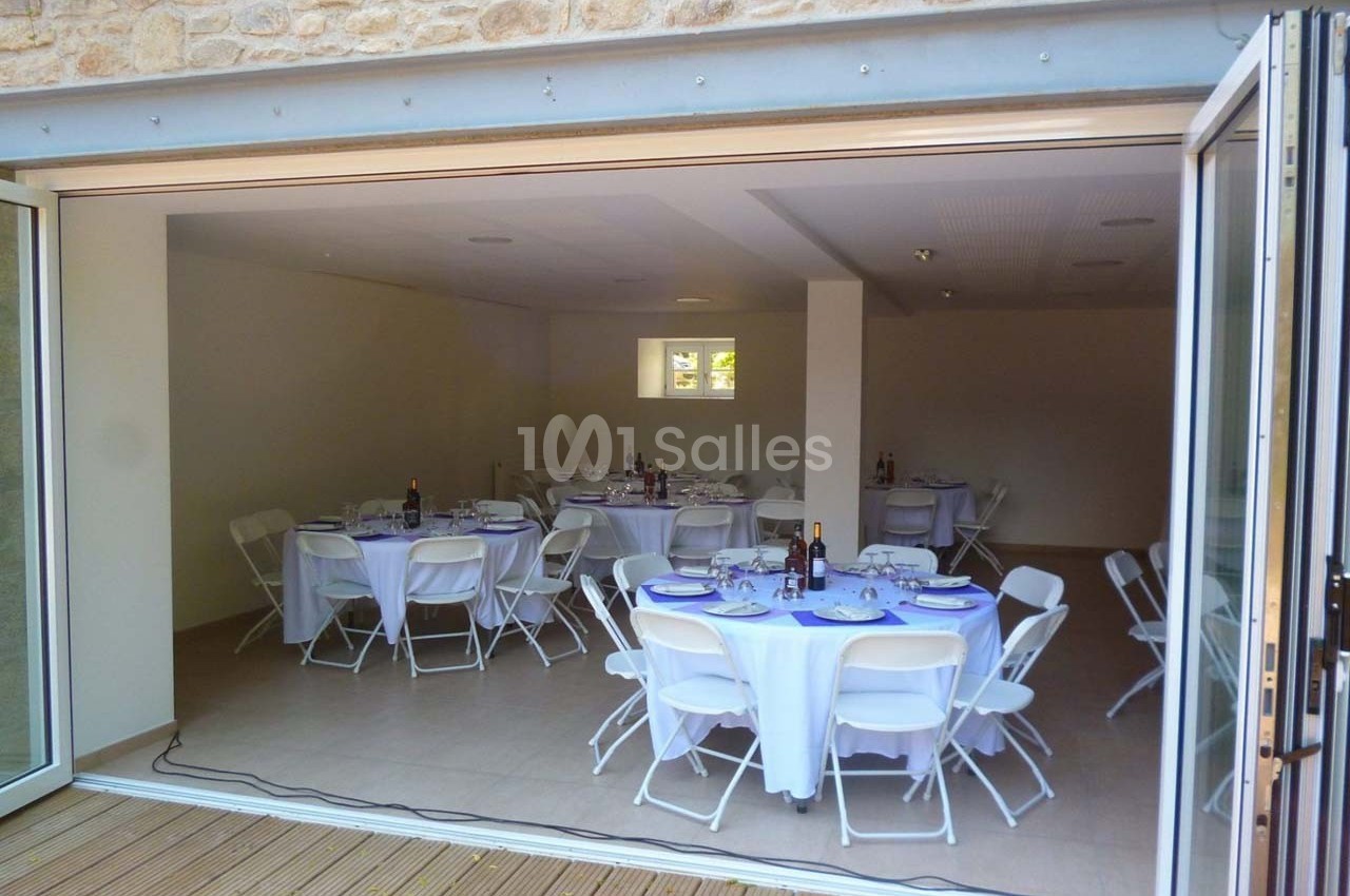 Salle de réception avec tables rondes dressées, chaises blanches et grandes portes vitrées ouvertes sur une terrasse en bois.