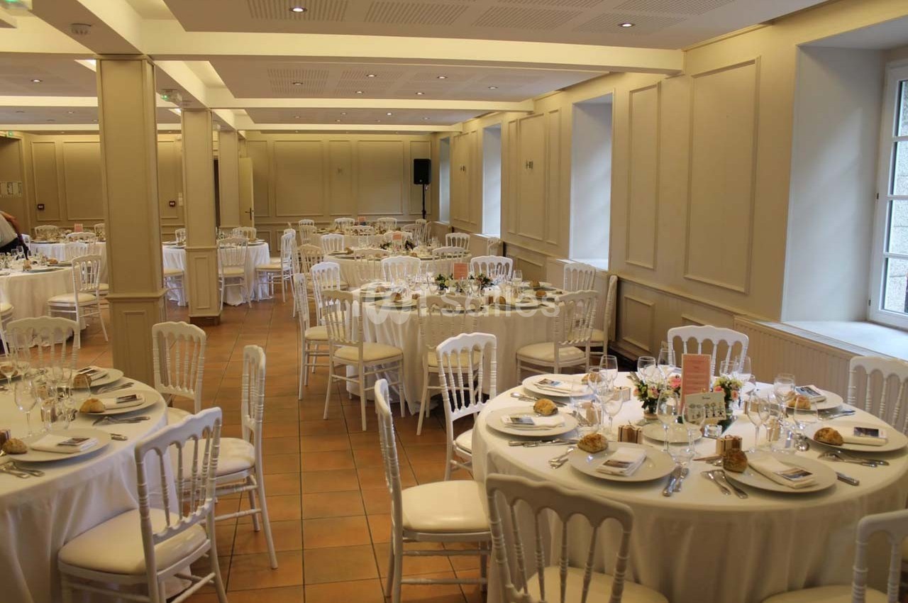 Salle de réception lumineuse avec des tables rondes dressées pour un repas, décorées de nappes blanches et de fleurs.