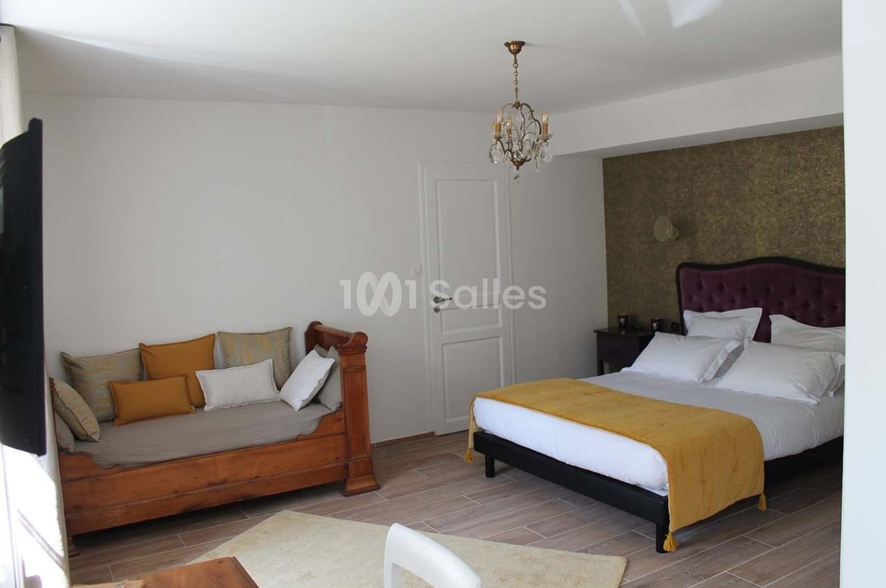 Chambre lumineuse avec un lit double, un canapé en bois, un lustre et des murs blancs, décorée dans des tons chauds.