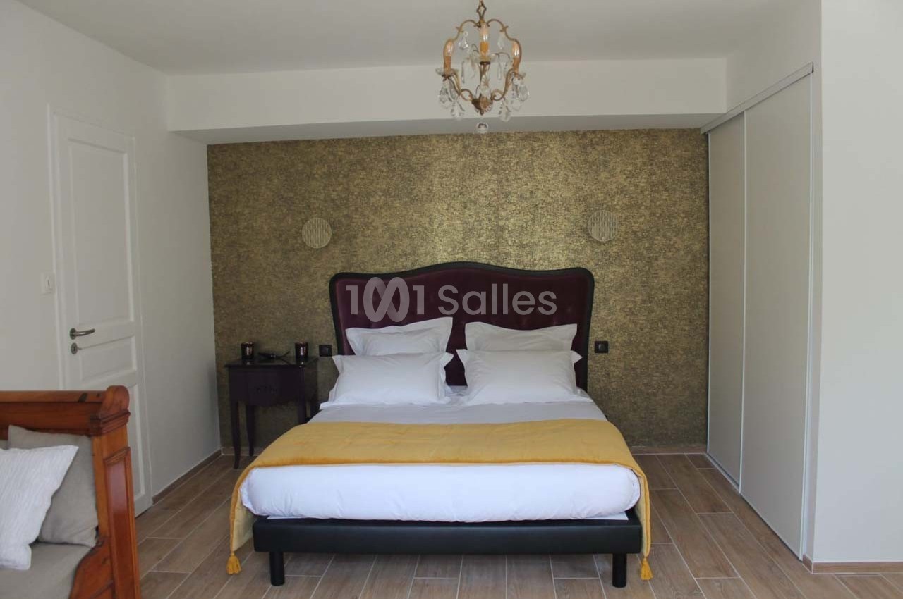 Lit double avec tête de lit capitonnée, draps blancs et couverture jaune, dans une chambre au mur doré texturé.