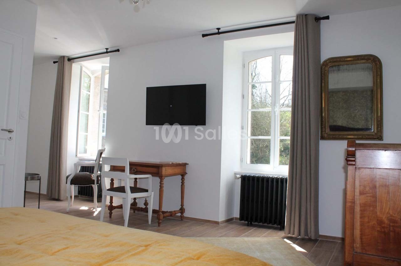 Chambre lumineuse avec lit, bureau en bois, télévision murale, fenêtres avec rideaux et radiateur noir.