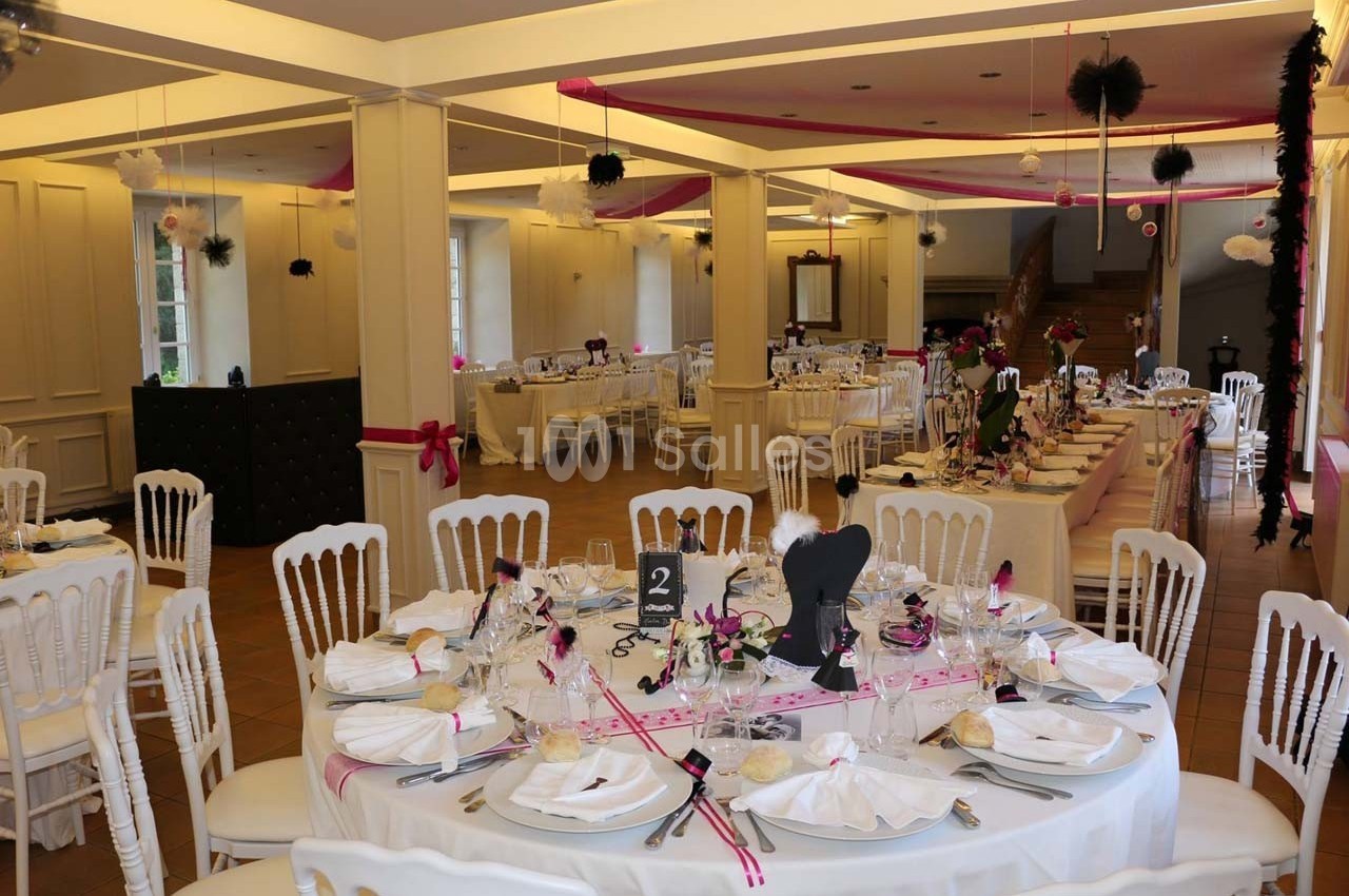 Salle de réception décorée avec des tables rondes et rectangulaires, nappes blanches et ornements roses et noirs.