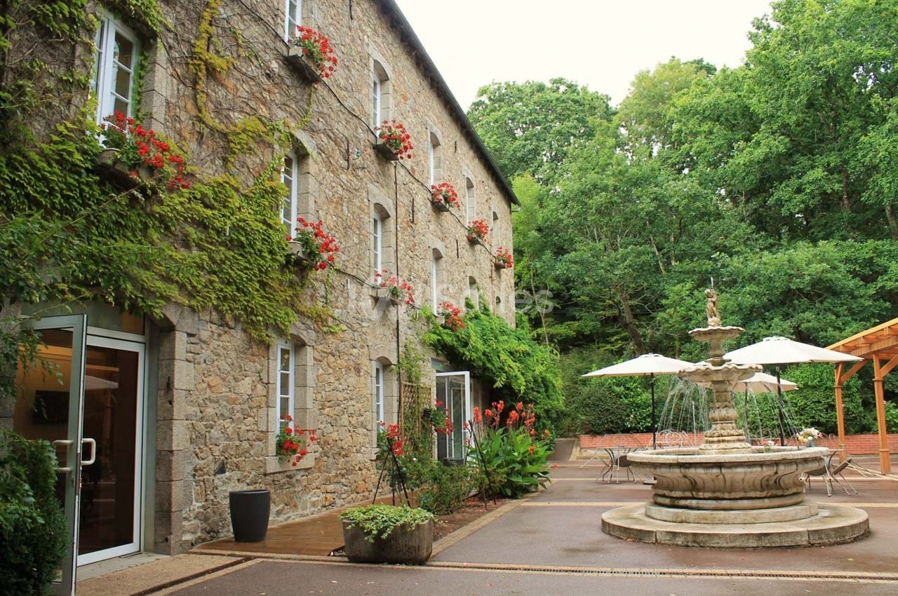 Façade en pierre d'un bâtiment avec des fenêtres fleuries, une fontaine centrale et une terrasse entourée de verdure.