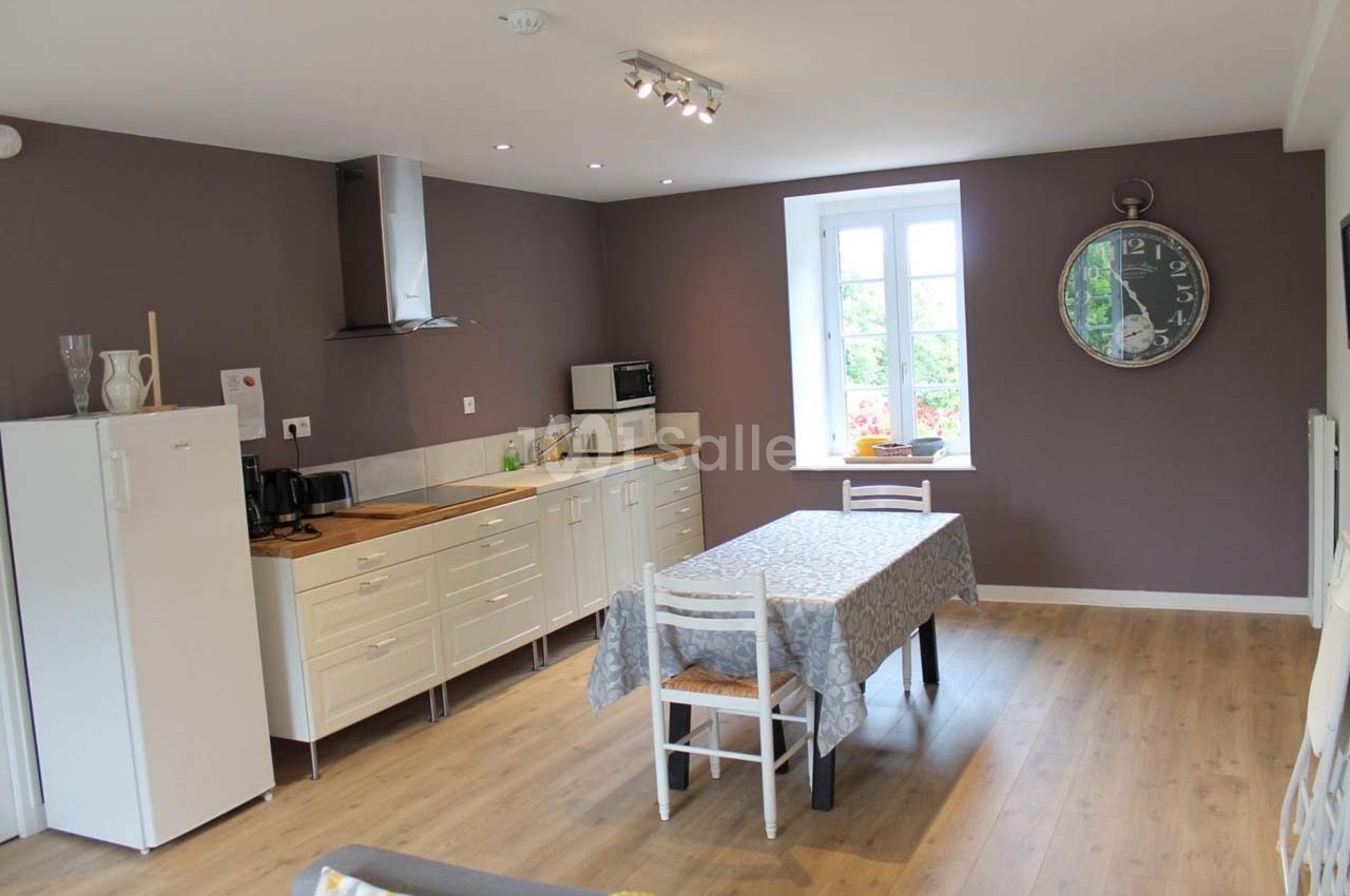 Cuisine lumineuse avec murs violets, meubles blancs, table avec nappe et grande horloge murale.