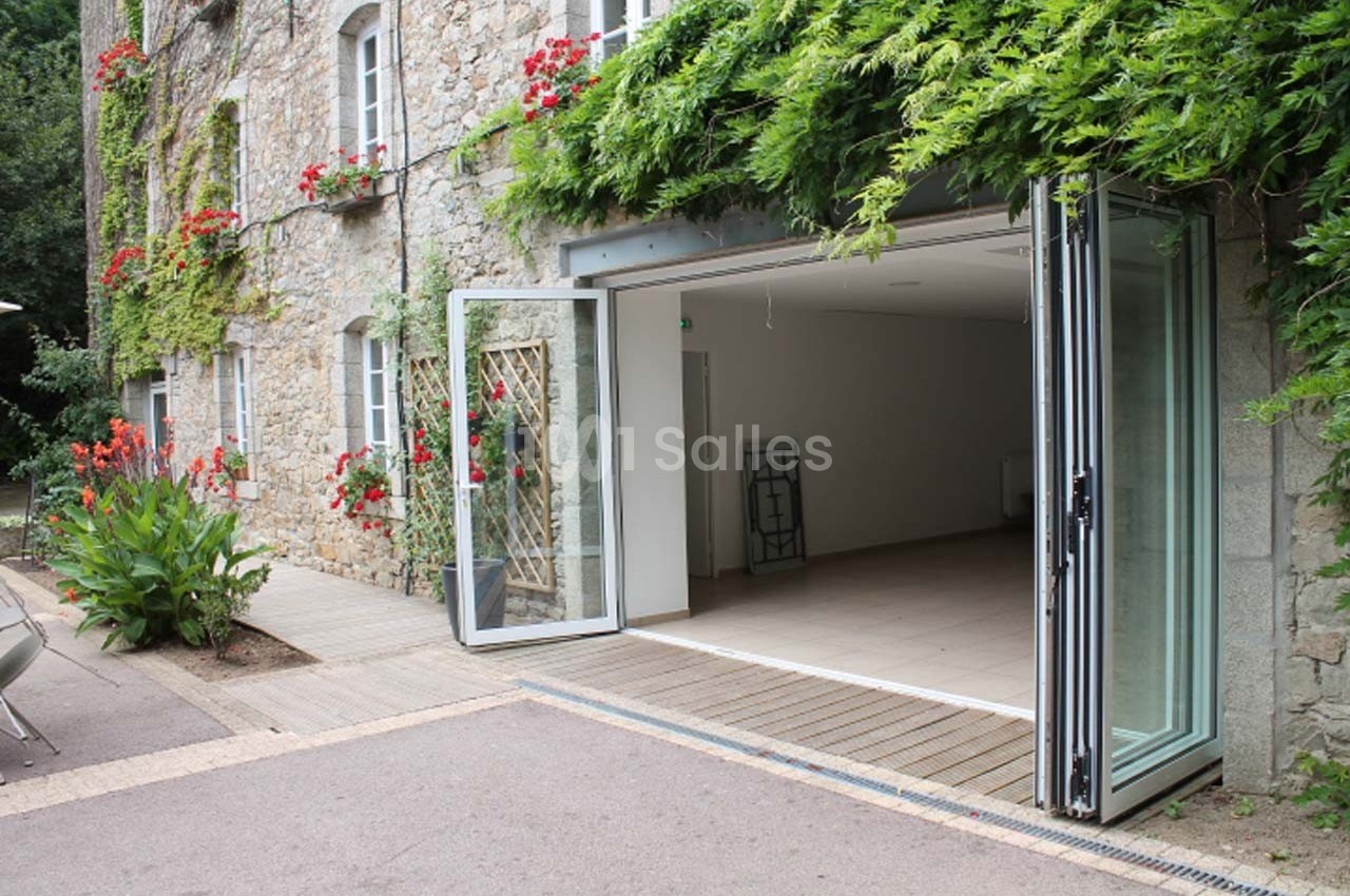 Façade en pierre avec porte vitrée pliable ouverte sur un intérieur vide, entourée de végétation et de fleurs rouges.