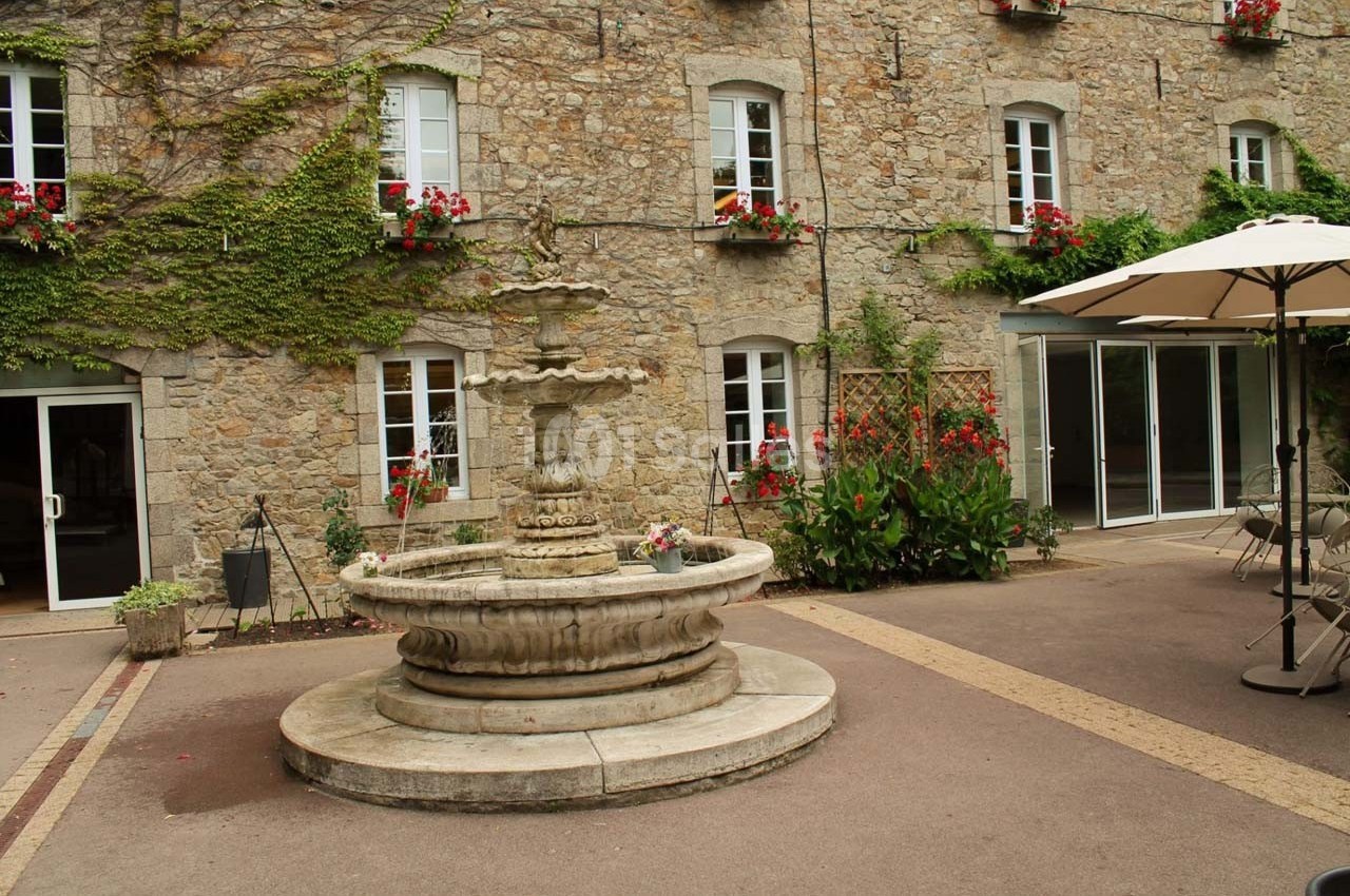 Cour intérieure avec une fontaine en pierre au centre, entourée de murs en pierre avec des fenêtres et des fleurs.