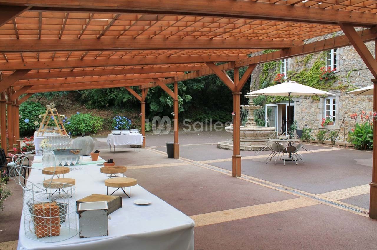 Espace extérieur avec pergola en bois, tables dressées, fontaine en pierre et végétation en arrière-plan.