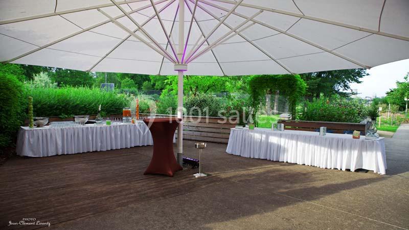 Grand parasol blanc abritant des tables drapées de nappes blanches dans un espace extérieur verdoyant.