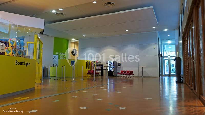Hall d'entrée lumineux avec une boutique à gauche, des sièges rouges au fond et une porte vitrée à droite.
