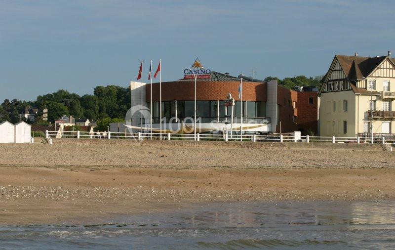 Façade d'un casino moderne en bord de plage, entouré de bâtiments traditionnels et de verdure.