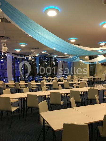 Salle décorée avec des guirlandes bleues et blanches, tables et chaises alignées, ambiance lumineuse et festive.