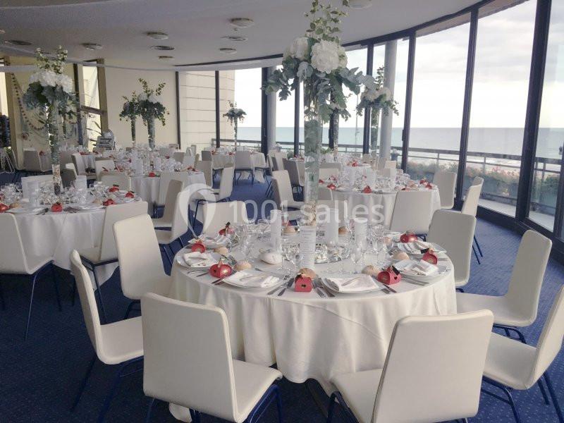 Salle de réception lumineuse avec des tables rondes dressées, décorations florales et vue sur la mer.