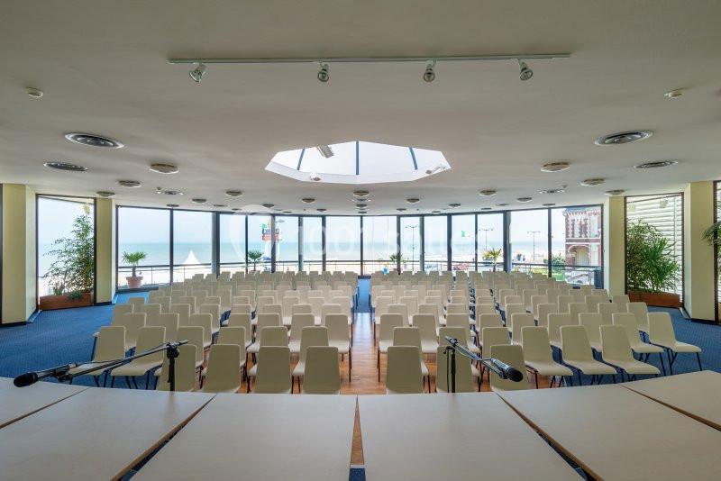 Salle de conférence lumineuse avec des rangées de chaises blanches, vue sur la mer à travers de grandes baies vitrées.