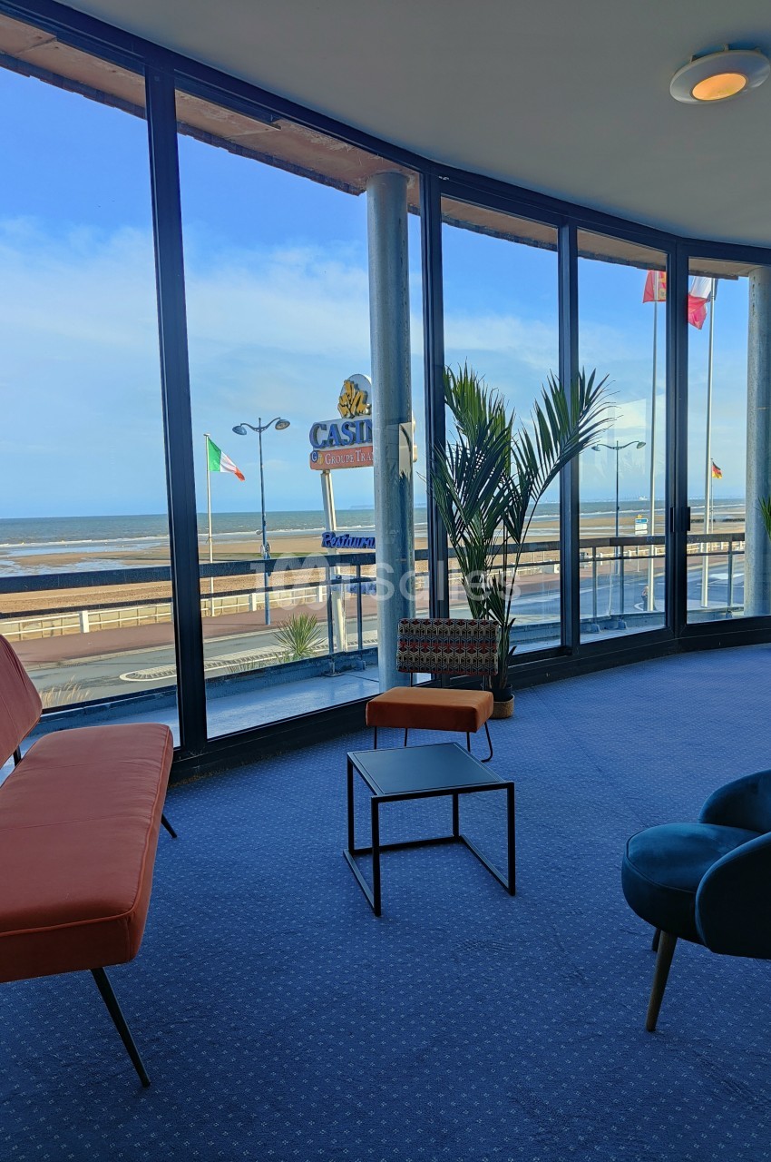 Vue sur la plage et la promenade depuis une salle lumineuse avec des fauteuils et de grandes baies vitrées.