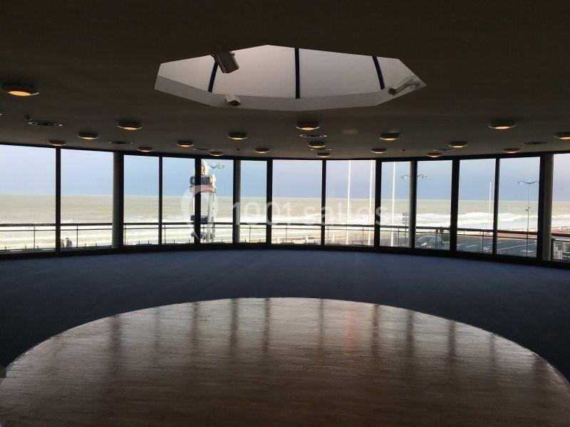 Salle circulaire avec de grandes baies vitrées offrant une vue sur la mer et une promenade en bord de plage.