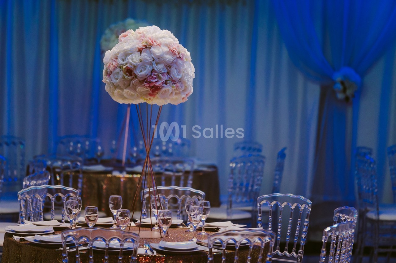 Centre de table floral avec des roses blanches et roses, entouré de chaises transparentes dans une salle décorée.