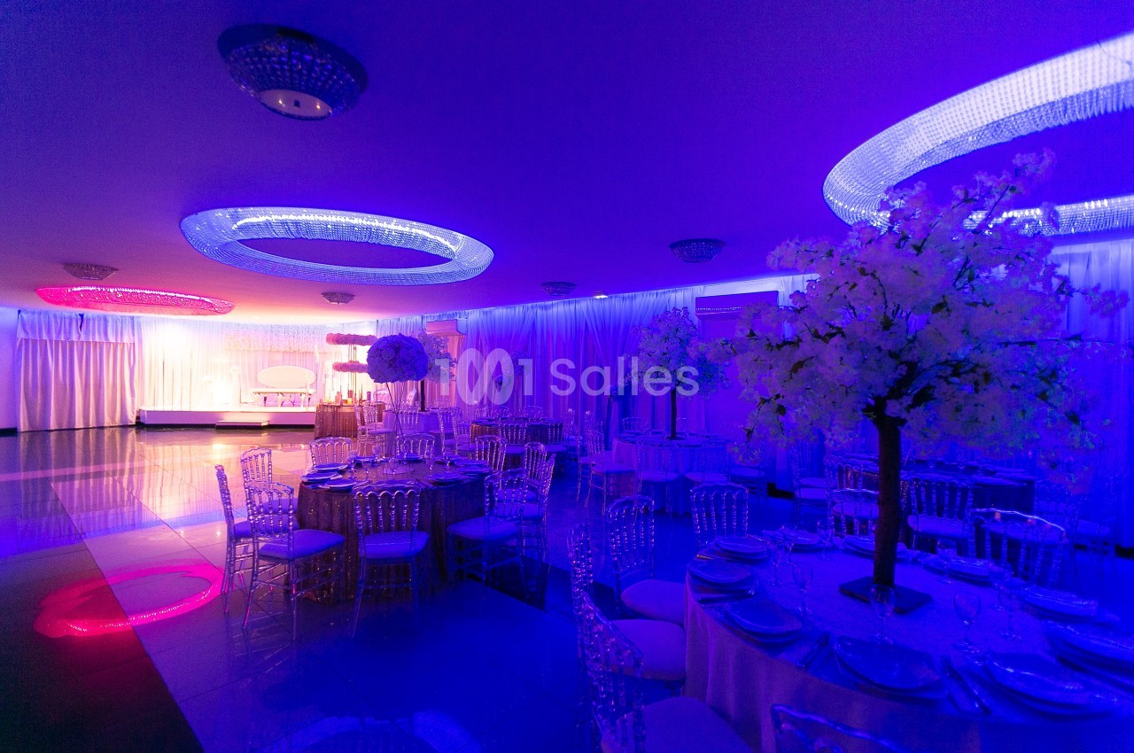 Salle de réception décorée avec des tables dressées, éclairages colorés et arbres artificiels ornés de fleurs blanches.
