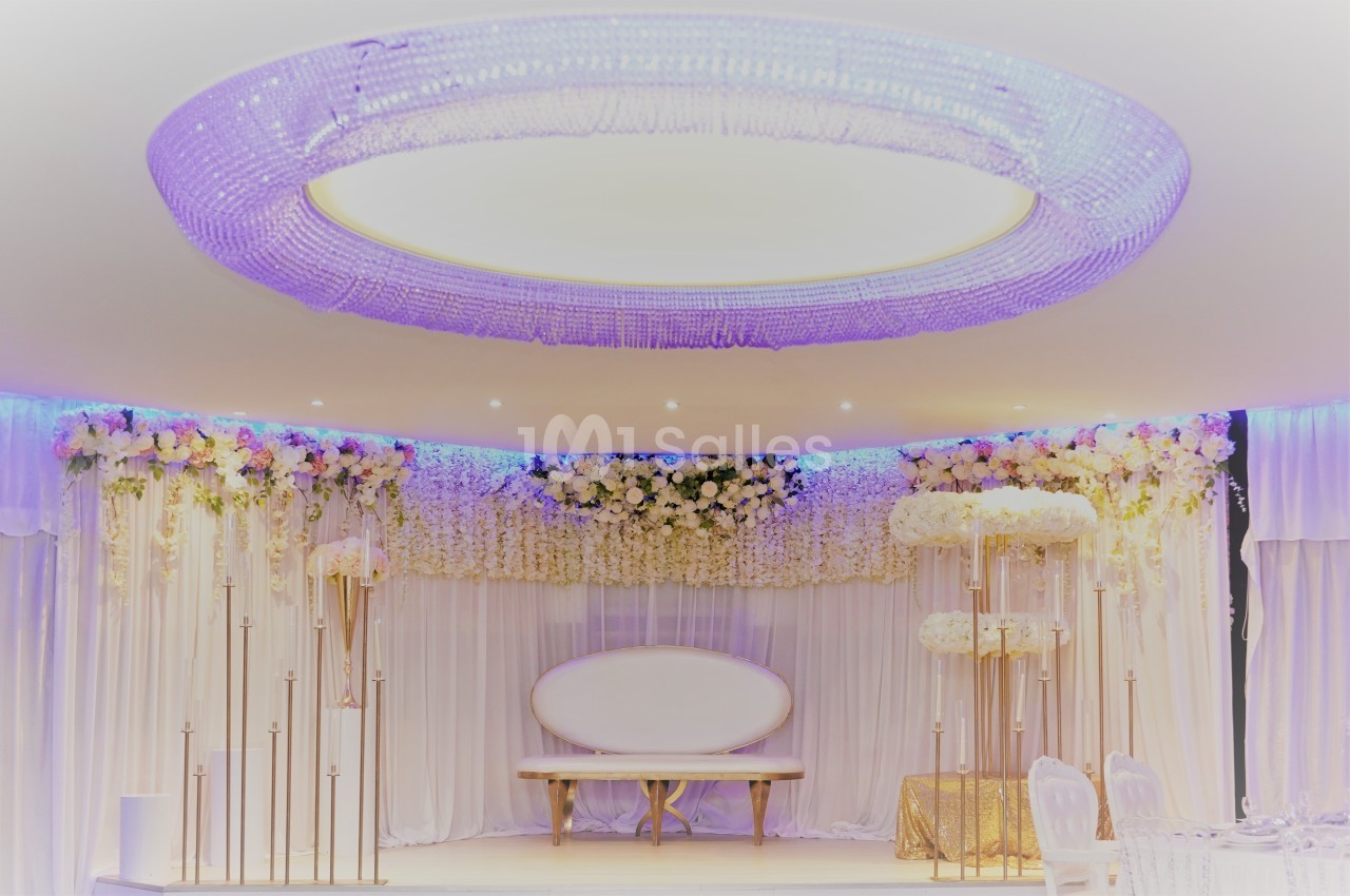 Décor de salle de réception avec un canapé blanc, rideaux et guirlandes de fleurs blanches sous un lustre circulaire…