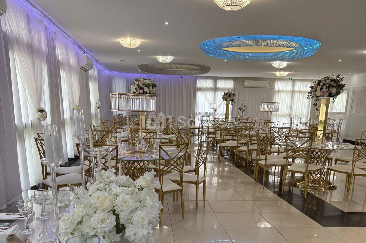Salle de réception élégante avec tables décorées, chaises dorées et arrangements floraux blancs sous un éclairage moderne.
