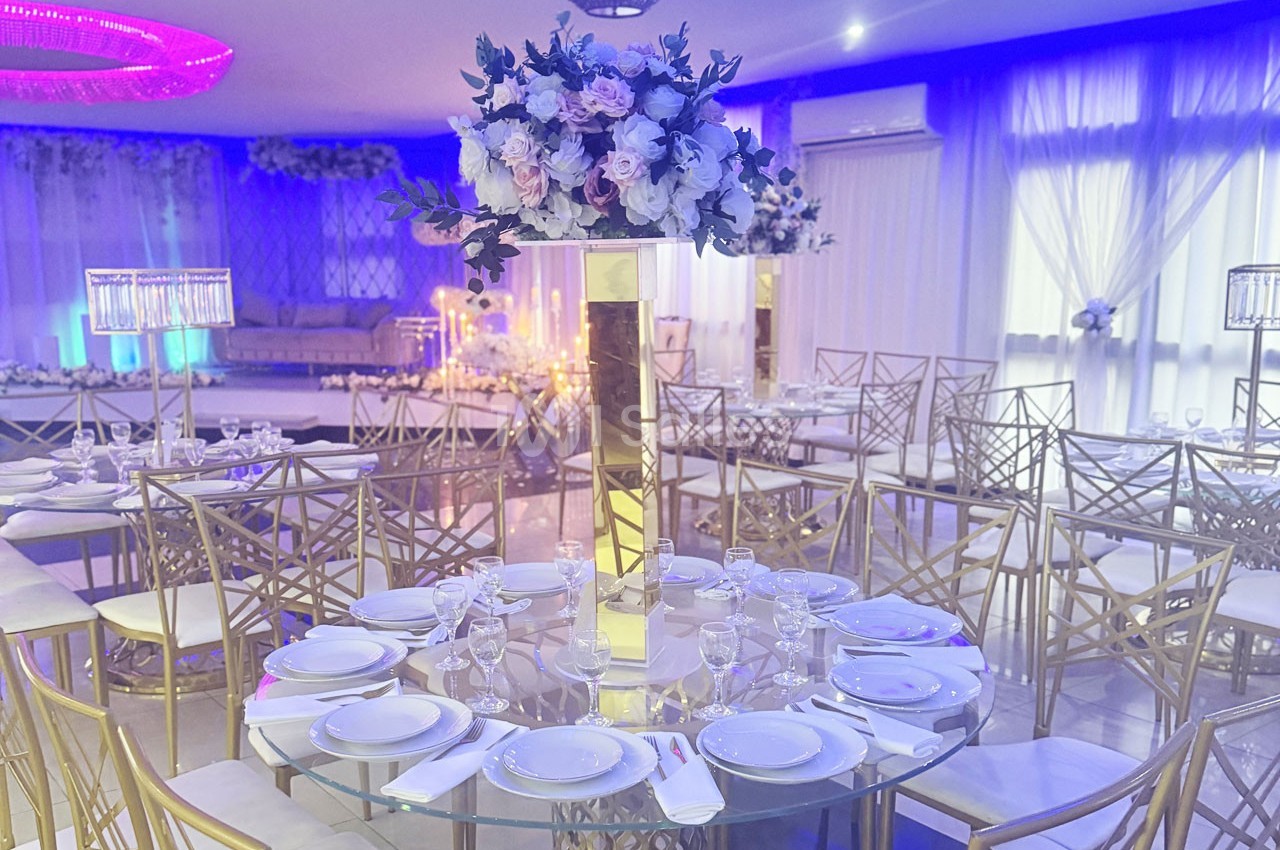 Salle de réception élégante avec tables rondes dressées, décor floral central et éclairage bleu tamisé.