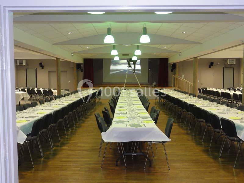 Salle de réception avec deux longues rangées de tables dressées, éclairée par des suspensions et équipée d'un écran au fond.