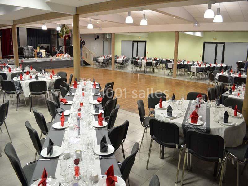 Salle de réception aménagée avec des tables dressées, nappes blanches et serviettes rouges, prête pour un événement.