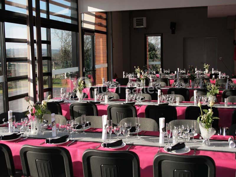 Salle de réception aménagée avec des tables dressées, nappes roses, vaisselle et décorations florales.