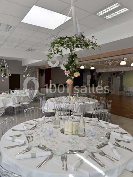 Salle de réception décorée avec des tables rondes dressées, chaises transparentes et suspensions florales.