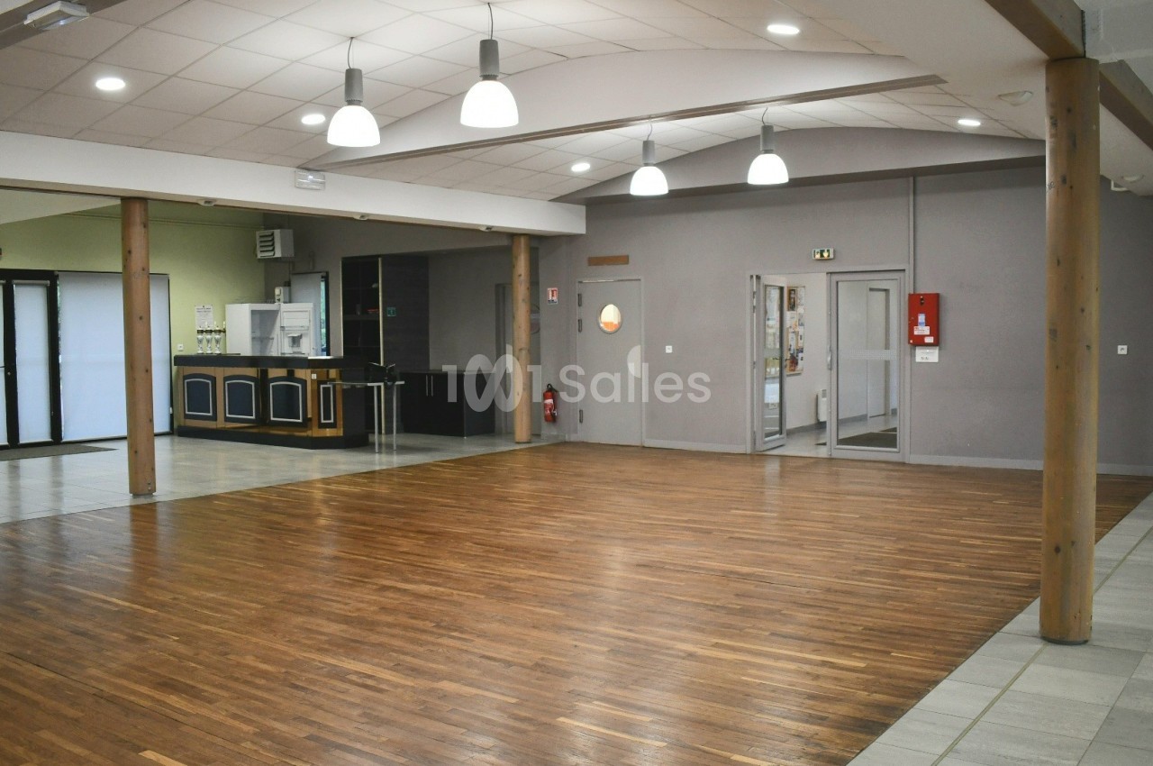 Salle polyvalente vide avec parquet, murs clairs, éclairage suspendu et coin cuisine en arrière-plan.