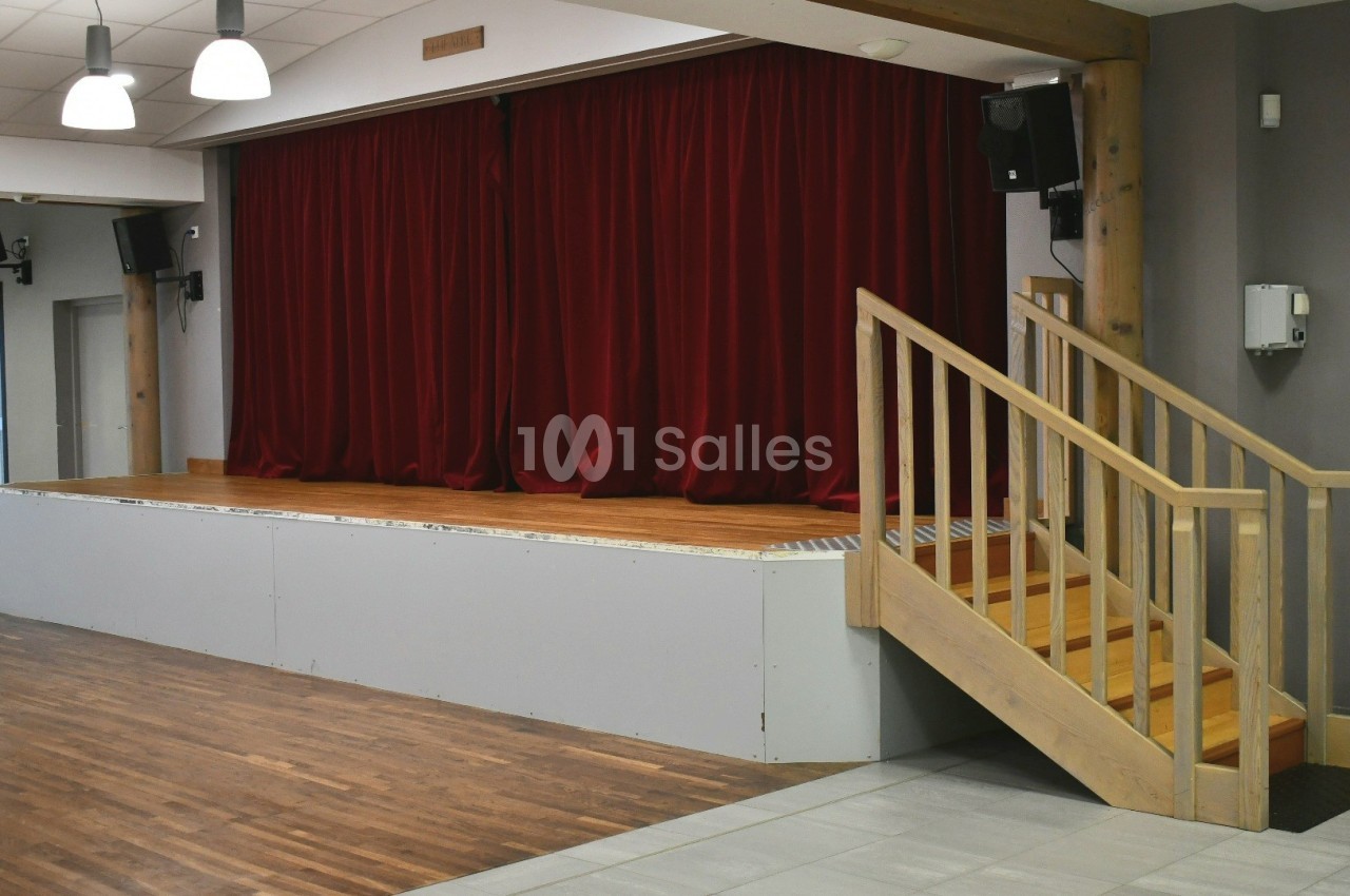 Scène en bois avec rideaux rouges fermés, éclairée par des lampes suspendues, et escalier en bois sur le côté.