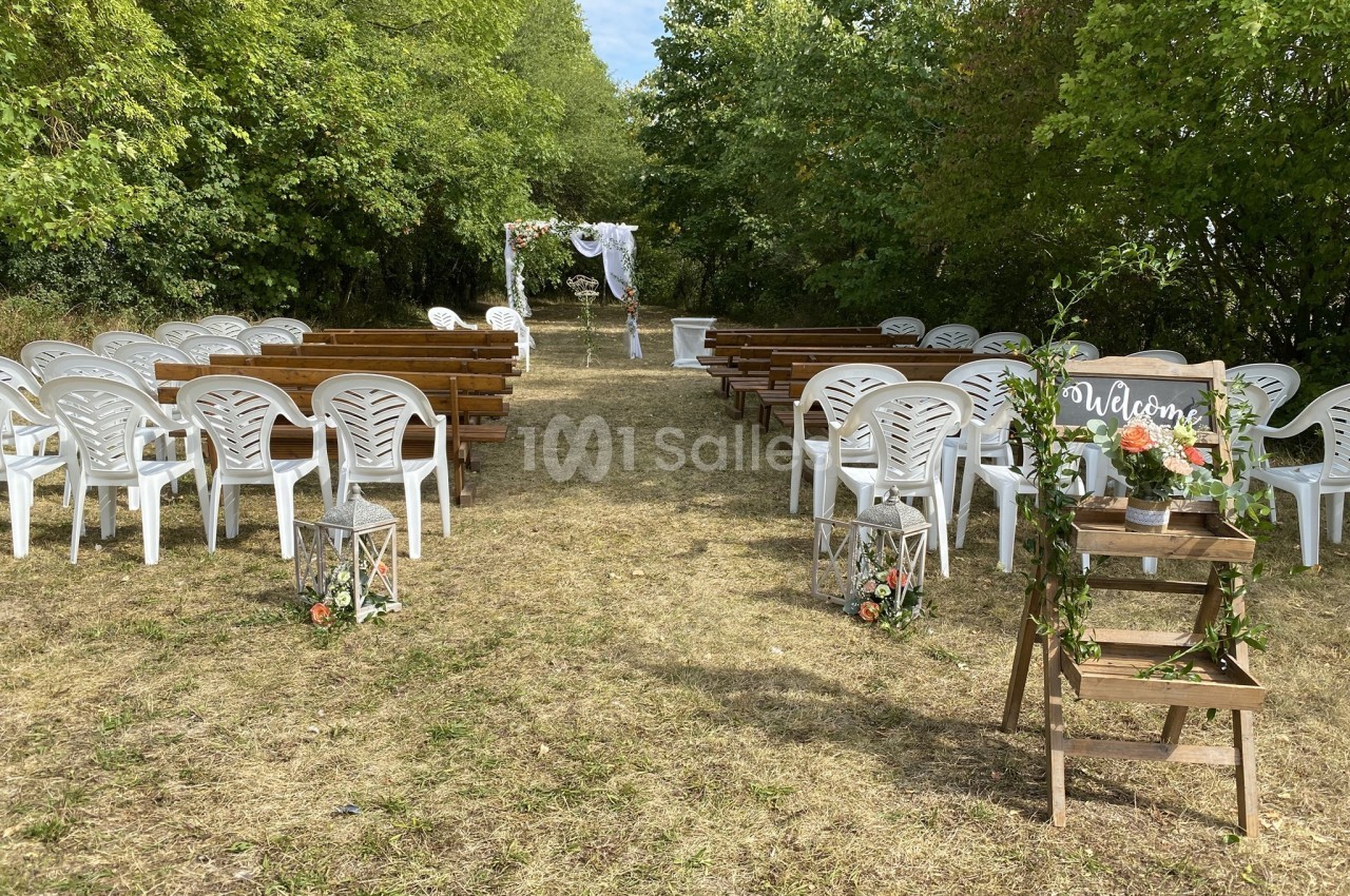 Allée centrale bordée de chaises blanches et bancs en bois menant à une arche de mariage décorée dans un cadre extérieur…