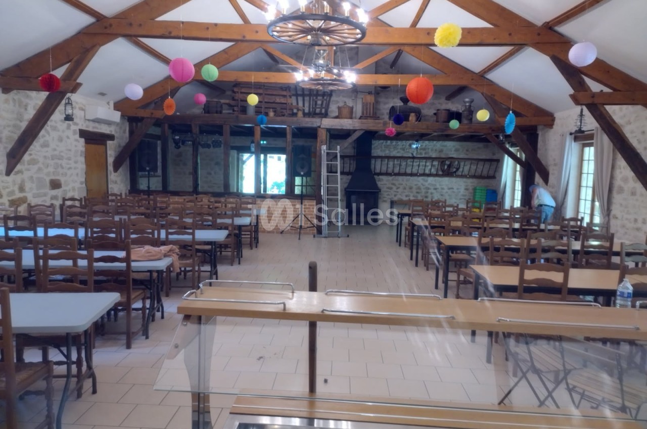 Salle de réception avec poutres apparentes, tables et chaises alignées, décorée de boules colorées suspendues.