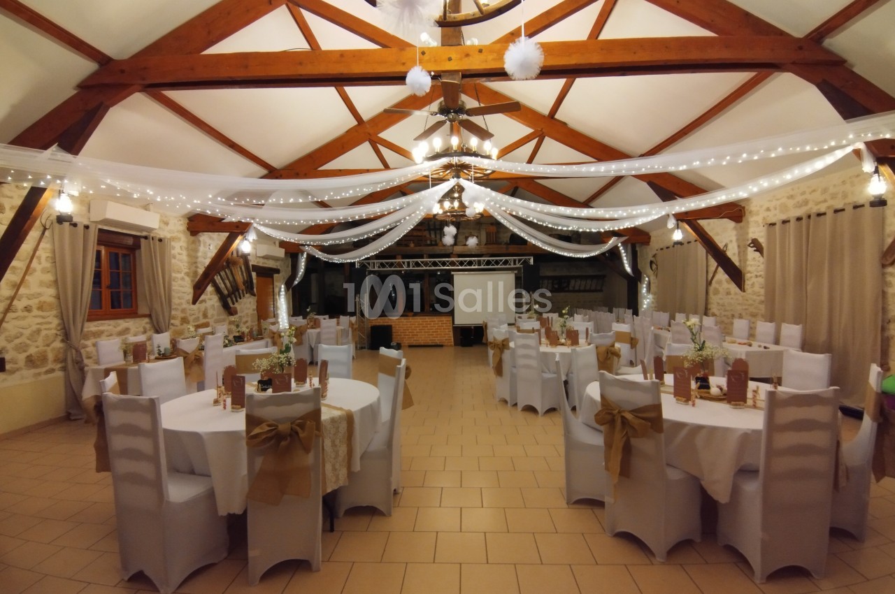 Salle de réception décorée avec des tables rondes, des chaises blanches, des guirlandes lumineuses et des poutres apparentes.