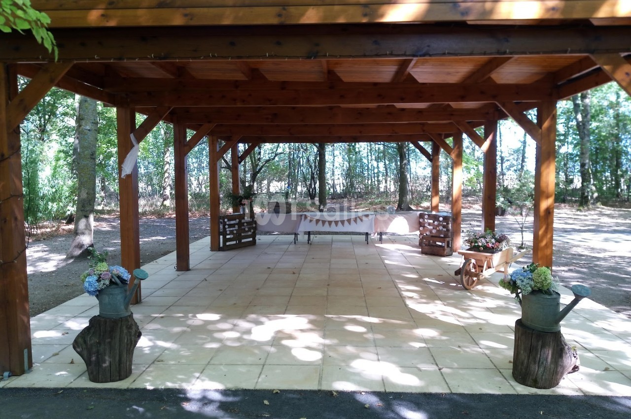 Structure en bois ouverte sur une terrasse carrelée, décorée de fleurs et d'éléments champêtres, entourée d'arbres.
