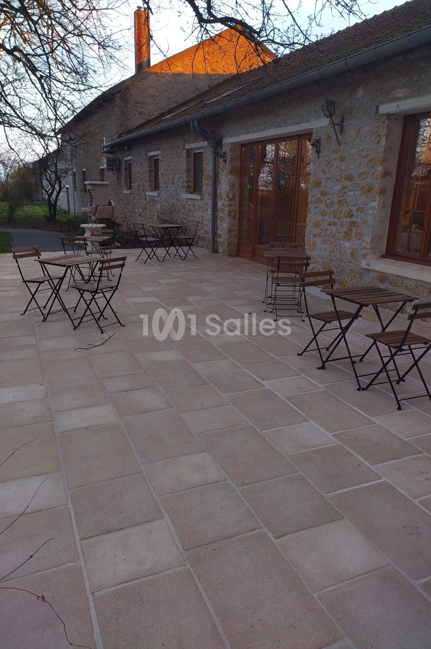 Terrasse en pierre avec tables et chaises en métal devant une maison en pierre, entourée d'arbres au coucher du soleil.
