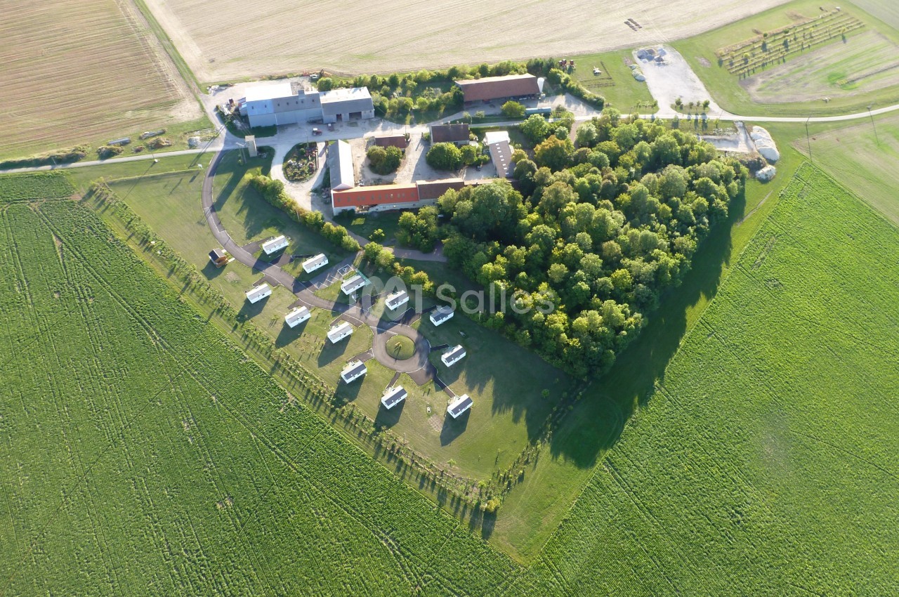 Vue aérienne d'un complexe avec bâtiments, espaces verts, arbres et structures dispersées au milieu de champs agricoles.