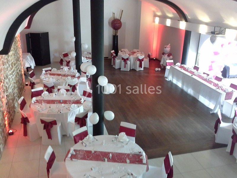 Salle de réception décorée pour un événement, avec tables rondes nappées de blanc et ornées de rubans rouges.
