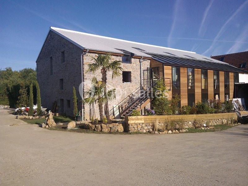 Bâtiment en pierre rénové avec une extension moderne en bois et verre, entouré de végétation et d'une cour dégagée.