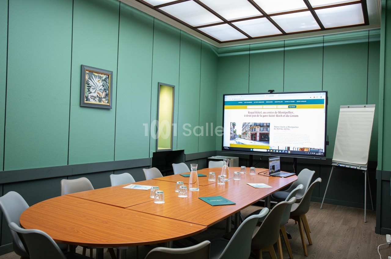 Salle de réunion avec table ovale en bois, chaises grises, écran allumé, tableau blanc et murs verts.